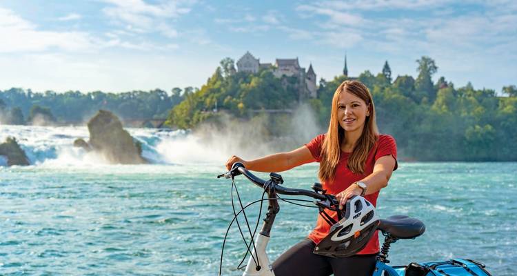 Mujer con una bicicleta frente a una cascada y un castillo.