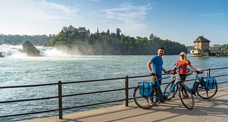 Pareja con bicicletas frente a un paisaje de río y castillo.