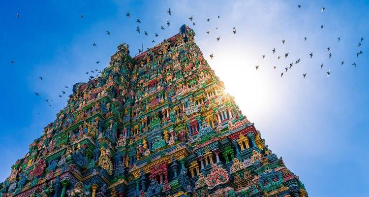 Temple spire with a flock of birds mid-flight against a blue sky.