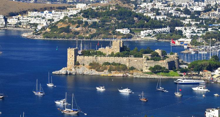 Castillo medieval dominando una pequeña península rodeada de un azul profundo mar salpicado de veleros