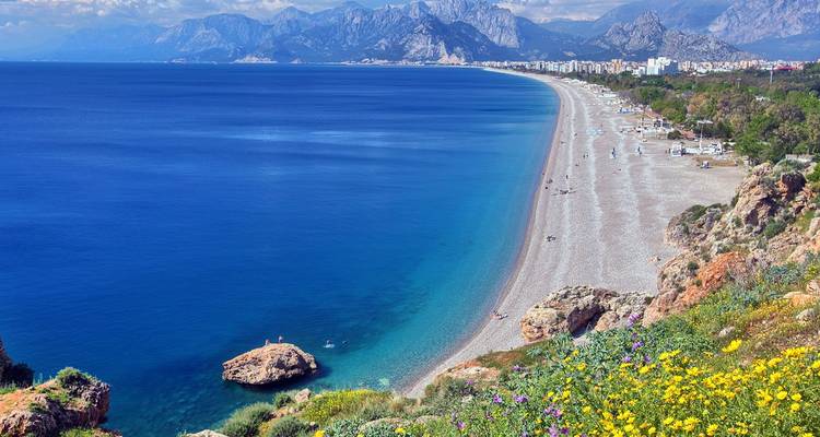 Expansive pebble beach with vivid blue Mediterranean waters and dramatic mountain backdrop near Antalya.