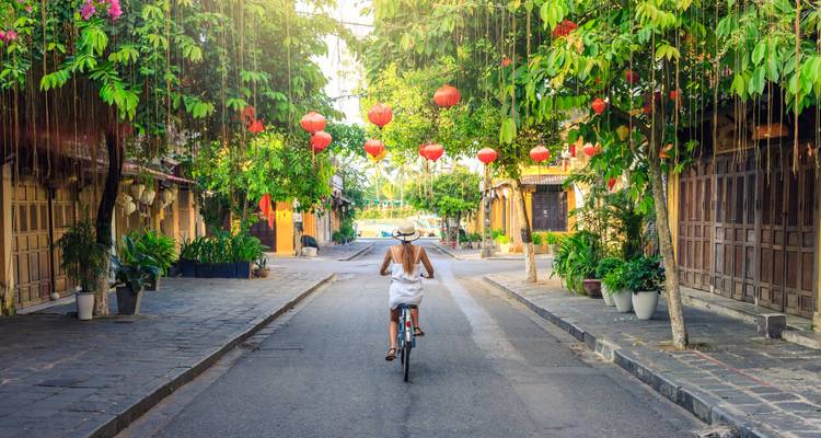Woman in white dress cycling down a lantern-adorned heritage street in Hoi An at golden hour.