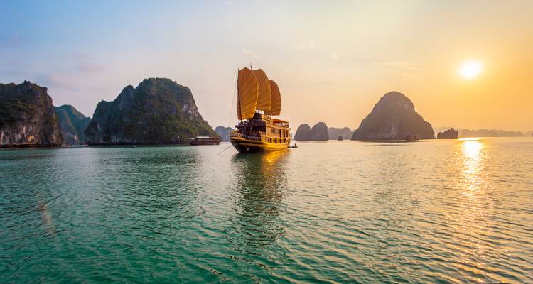 Traditional junk boat sailing between limestone karsts on Halong Bay during a golden sunset.