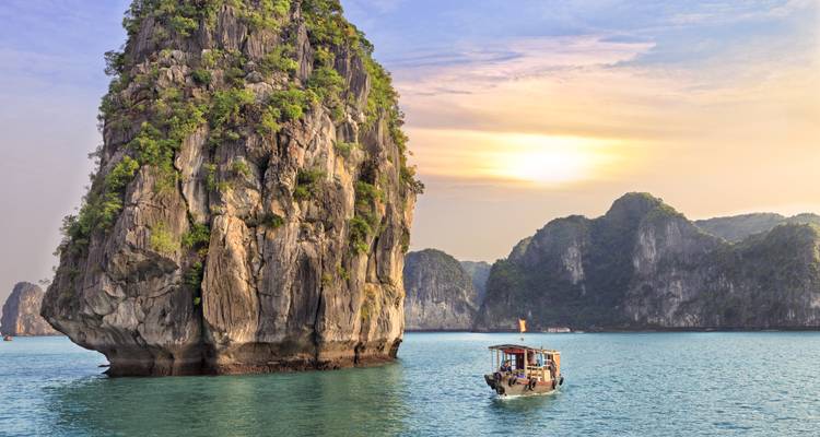 Small fishing boat dwarfed by towering limestone pillar against pastel sunset in Halong Bay.