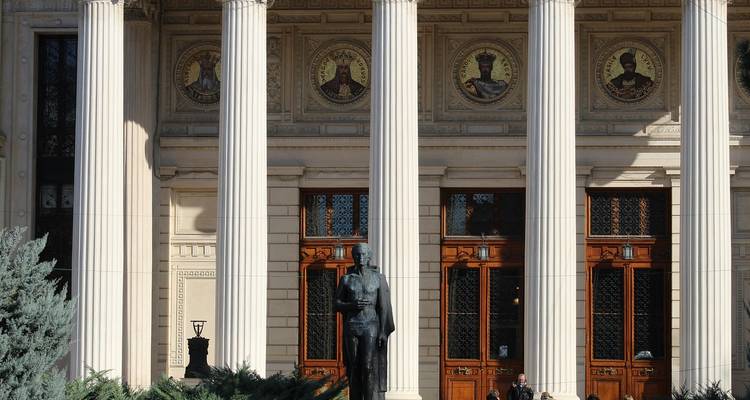 Statue devant un bâtiment néoclassique avec des colonnes et des portraits décoratifs.