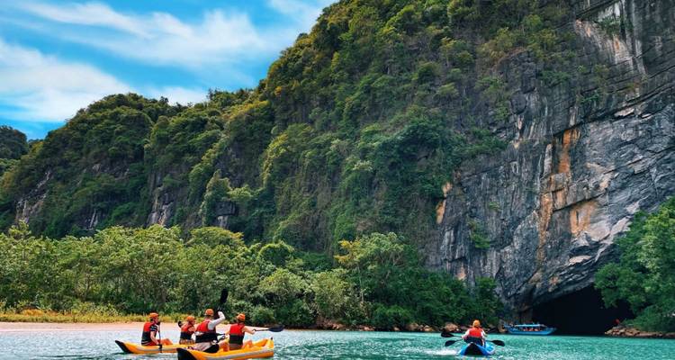 Tourists in kayaks rowing near a lush, forested mountainside with a cave in view.