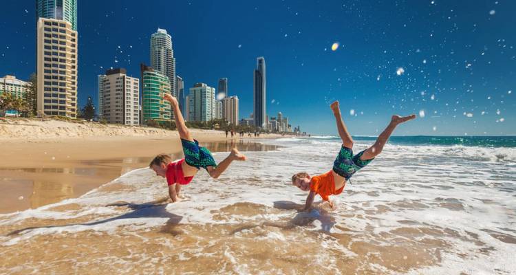 Two children do cartwheels in shallow surf on sunny beach with Gold Coast skyscrapers behind.