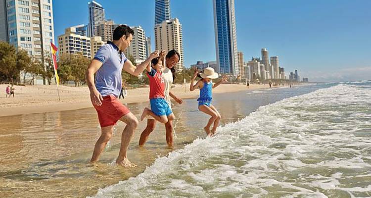 Family in swimwear hold hands and splash through waves along city beach backed by tall towers.