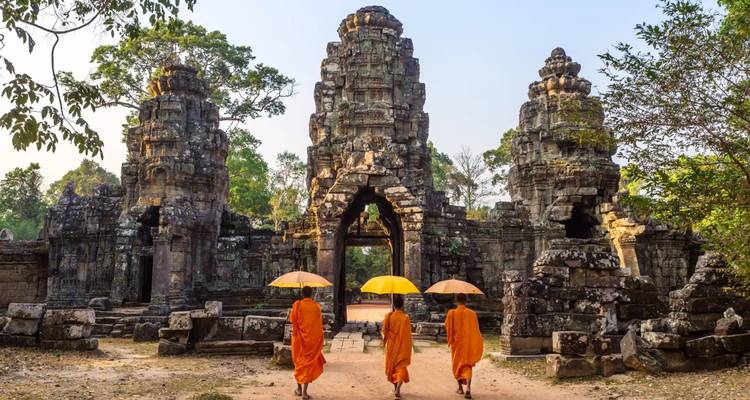 Monks in orange robes with umbrellas walking toward a stone temple with intricate carvings.