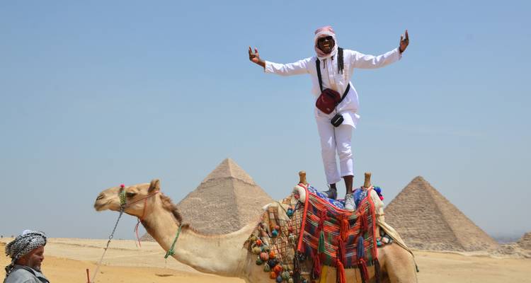 Man standing on a camel with the pyramids of Giza in the background.