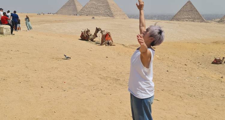 A person raising arms in front of the pyramids with camels in the background.
