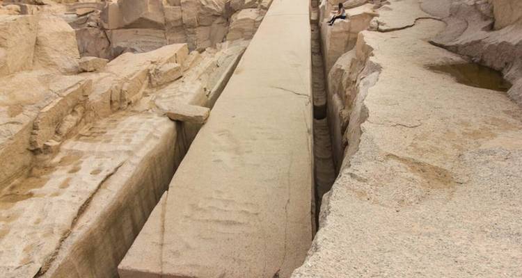 Obélisque massif inachevé gisant dans une carrière de granit avec un visiteur perché sur le bord pour donner l'échelle