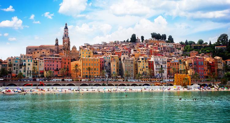 Colorful Mediterranean coastal town with swimmers enjoying a calm bay beneath terraced houses.