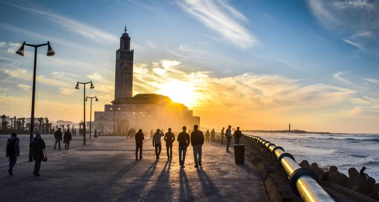 Peatones recortados en la pasarela de Casablanca al atardecer dorado con la Mezquita Hassan II en la niebla.