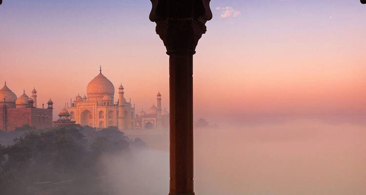 Dreamy sunrise view of the Taj Mahal emerging above mist, framed by a dark foreground pillar.