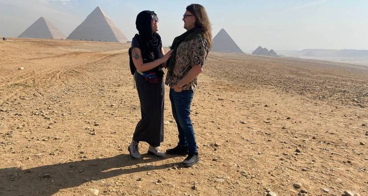 Pareja de pie en el desierto con pirámides al fondo.