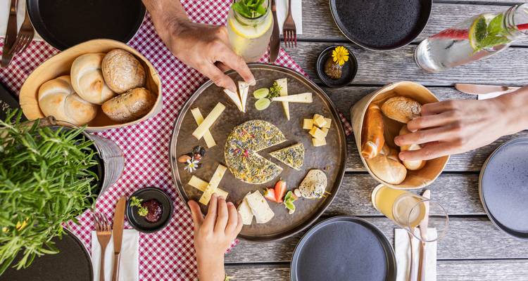 Hände, die nach verschiedenen Käsesorten und Brot auf einem Holztisch greifen.
