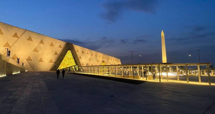 Extérieur nocturne du Grand Musée égyptien illuminé de lumières à côté d'un haut obélisque