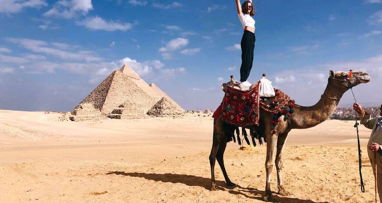 Femme en équilibre sur une selle de chameau dans le désert avec les pyramides de Giza en arrière-plan.