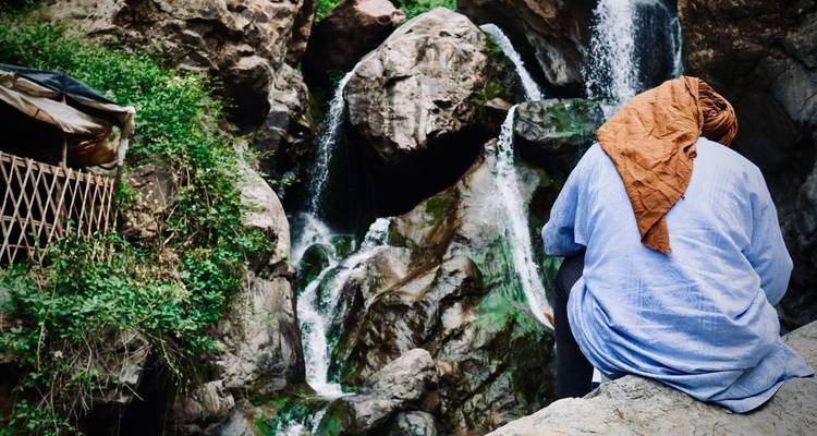 Persona sentada en rocas frente a una cascada en una zona montañosa.
