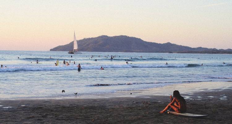Plage avec des surfeurs et un voilier au loin au coucher du soleil.