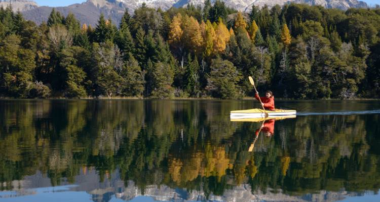 Kayakiste sur un lac tranquille avec des reflets d'arbres et de montagnes enneigées.