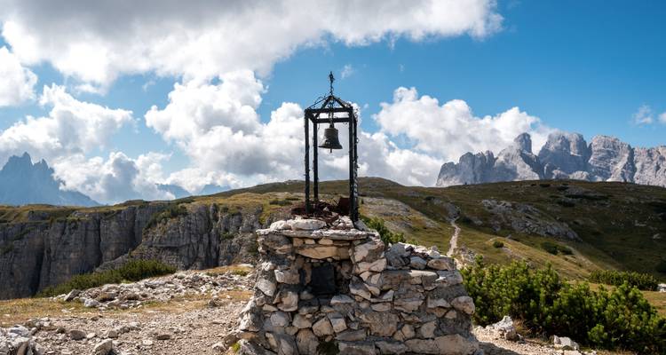 Une petite structure avec une cloche sur une colline rocheuse avec des montagnes en arrière-plan.