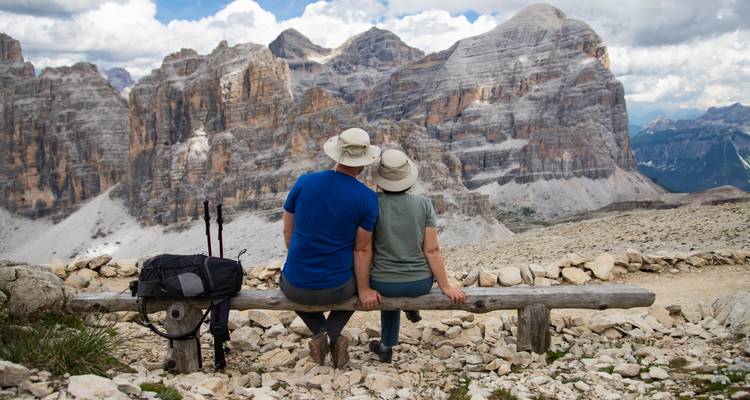 Couple assis sur un banc avec des bâtons de randonnée, surplombant un paysage montagneux.