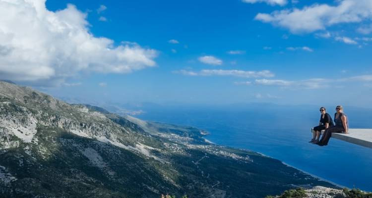 Zwei Personen, die auf einer Plattform sitzen mit einem weiten Blick auf Berge und Meer.