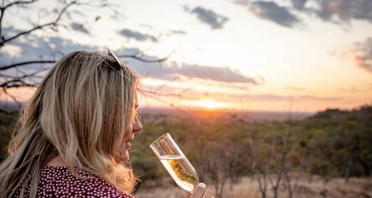 Woman enjoying a drink at sunset with greenery in the background.