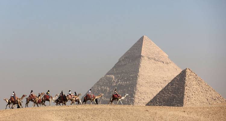 Group of people on camels with pyramids in the background.