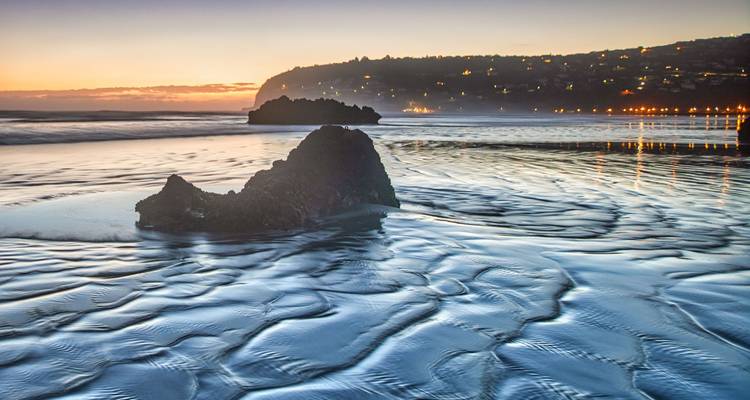 Una impresionante puesta de sol costera con patrones texturizados de arena que conducen a un afloramiento rocoso, con el mar y un pueblo en la distancia.