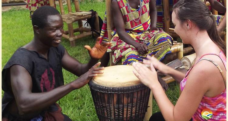 People playing drums in a grassy setting with traditional clothing and a wooden backdrop.