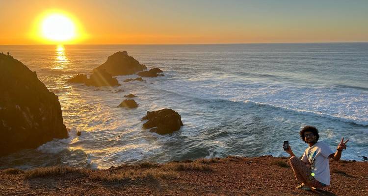 Une personne qui pose près d'une falaise avec le soleil qui se couche sur l'océan.