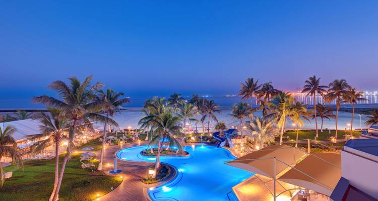 Nachtansicht eines Resorts mit beleuchtetem Pool und Palmen in Meeresnähe.