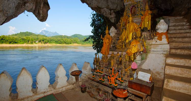 Ein mit goldenen Buddha-Statuen gefülltes Heiligtum in einer Flussuferhöhle mit Blick auf einen breiten Fluss und entfernte Hügel
