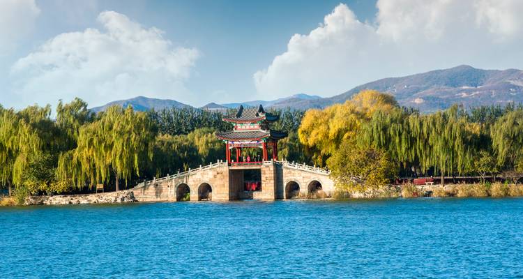 Traditional Chinese pavilion on a multi-arched bridge across a vivid blue lake with mountains behind.