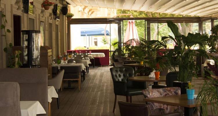 Café-Terrasse mit Sitzgelegenheiten und Pflanzen, teilweise überdacht.