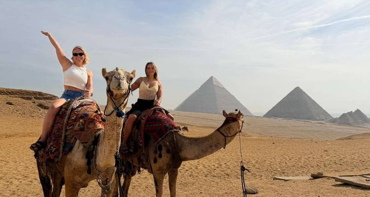 Zwei Frauen reiten auf Kamelen, während sich die Großen Pyramiden majestätisch hinter ihnen erheben.