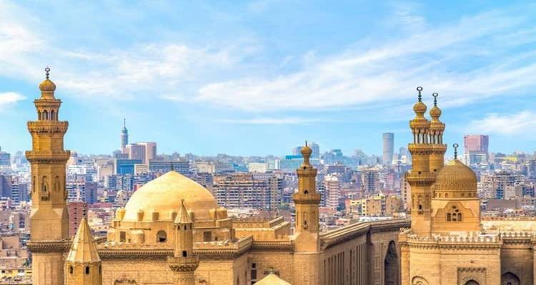 Panoramablick auf die Skyline von Kairo mit historischen Moscheekuppeln und Minaretten, die sich über die moderne Stadtlandschaft unter einem strahlend blauen Himmel erheben.