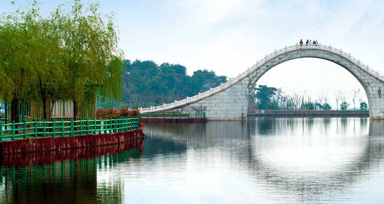 Puente arqueado sobre un lago tranquilo con árboles al fondo.