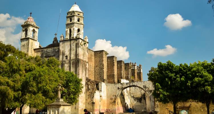 Muros de convento de piedra desgastada y campanarios enmarcados por árboles frondosos bajo un cielo azul con nubes.