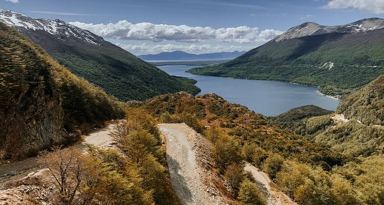 Punto de vista elevado sobre un lago de color zafiro rodeado de montañas empinadas cubiertas de bosque y una cordillera nevada a lo lejos.