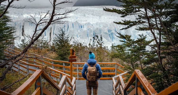 Un routard en veste bleue et bonnet de laine se tient sur des passerelles en bois face au mur de glace du glacier Perito Moreno.
