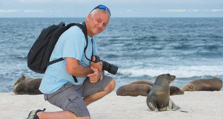 A man kneeling on a beach with sea lions and holding a camera.