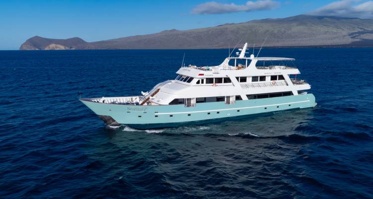 Luxury white and turquoise motor yacht cruising through deep blue Galápagos waters with volcanic islands in view.
