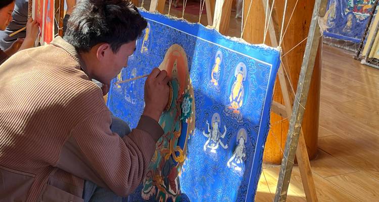 Un jeune artiste peignant avec concentration une thangka bleue à l'intérieur d'un studio aux planchers de bois