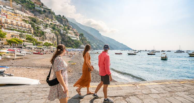 Three people walking near a beach with hills in the background.