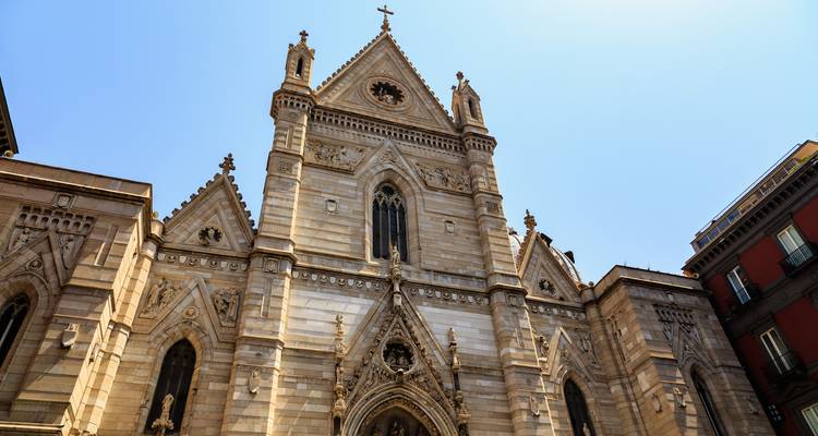 Sunlit façade of Naples Cathedral featuring ornate Gothic architecture and pointed spires against a clear sky.