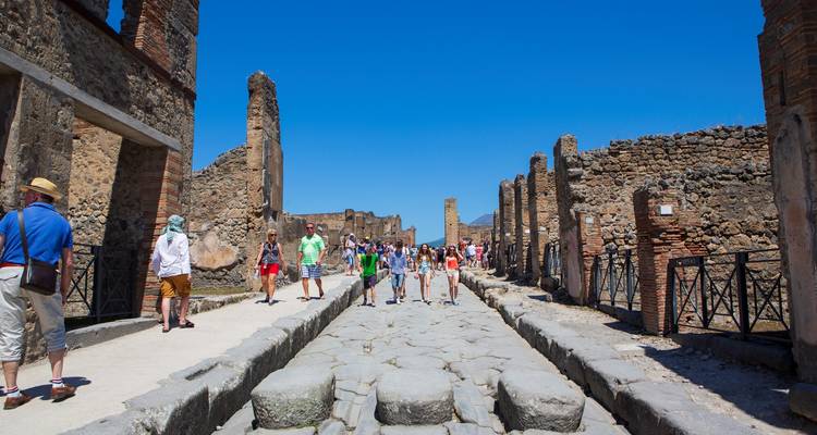 Tourists walking through a broad excavated street among the stone ruins of Pompeii on a clear day.